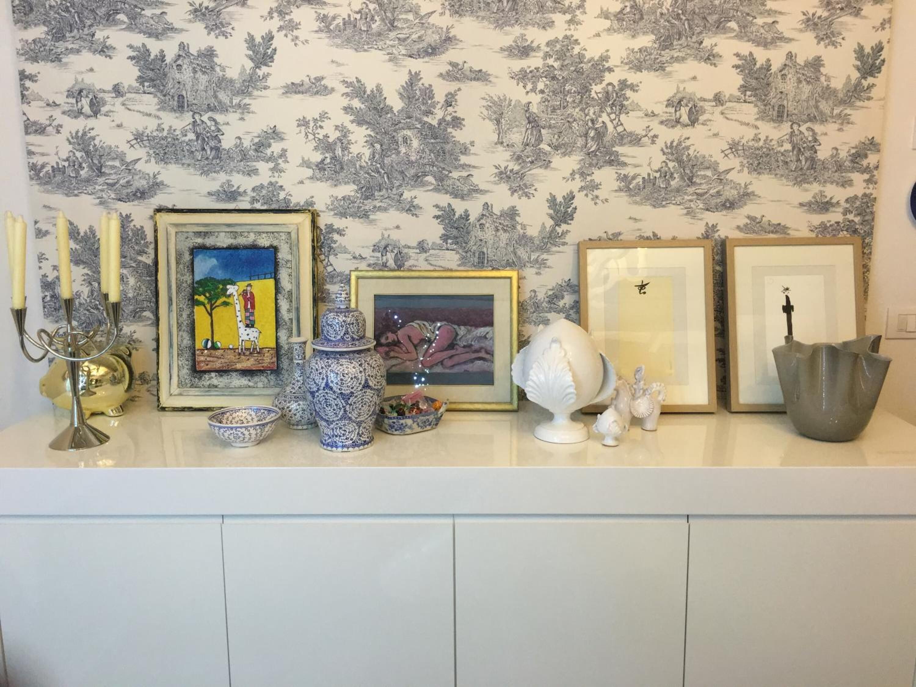 Pared con papel tapiz floral y una repisa con varios objetos decorativos, incluyendo cuadros enmarcados, un candelabro y objetos de cerámica.