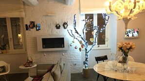 A cozy living room with a white brick wall, a flat-screen TV mounted above a fireplace, a dining table with chairs, and a sofa with a red and black checkered throw blanket. - Luxury My Home (Tolentino)