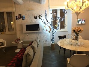 A cozy living room with a white brick wall, a flat-screen TV mounted above a fireplace, a dining table with chairs, and a sofa with a red and black checkered throw blanket. - Luxury My Home (Tolentino)