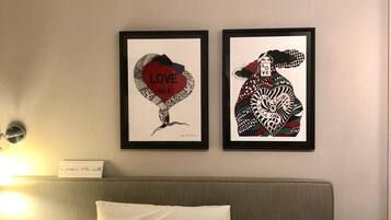 A hotel room with a bed, two framed artworks on the wall, a bedside table, and a wall-mounted light.