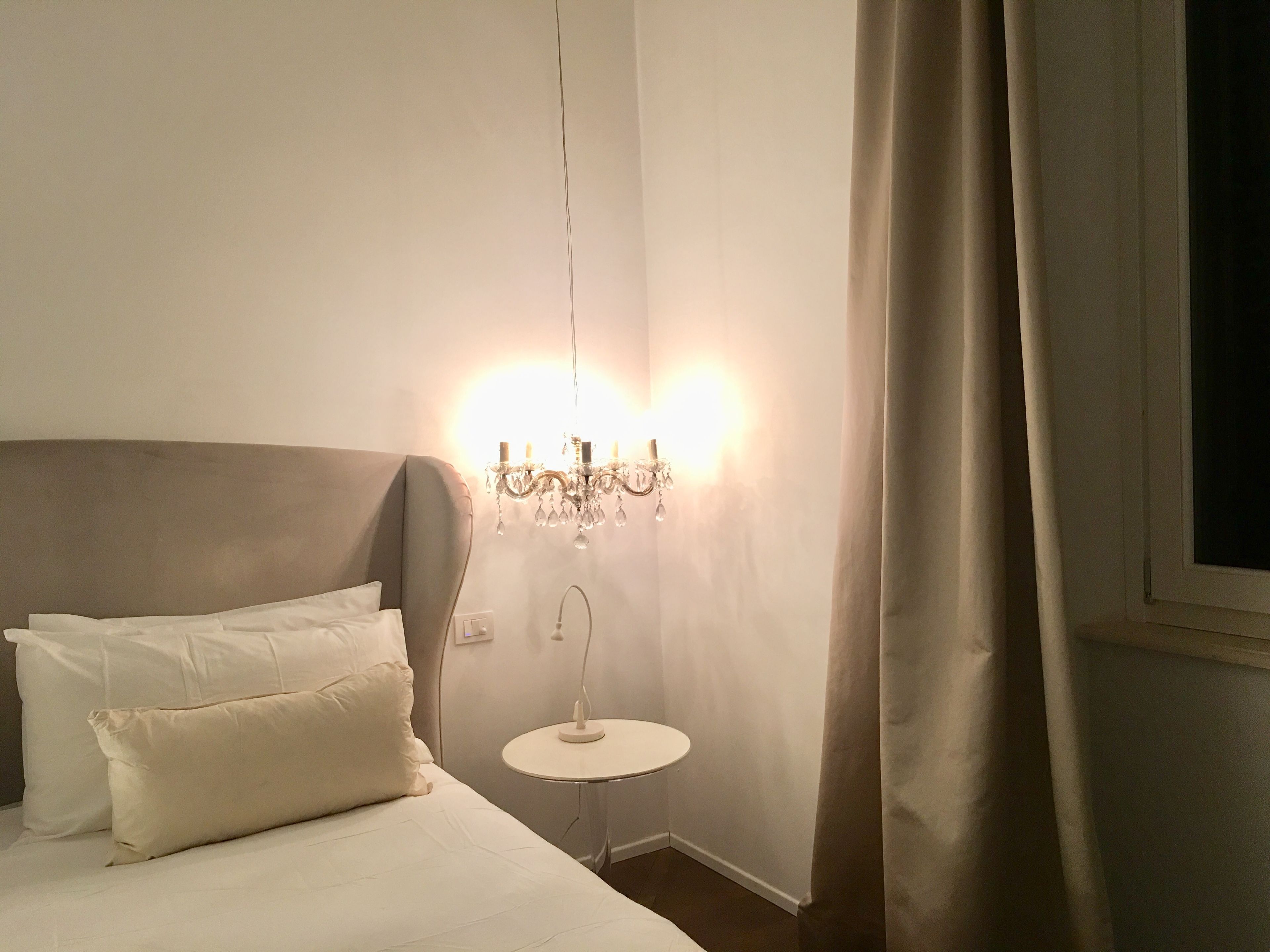 A neatly made bed with white linens and pillows, a small round table with a lamp, and a chandelier with multiple lights.