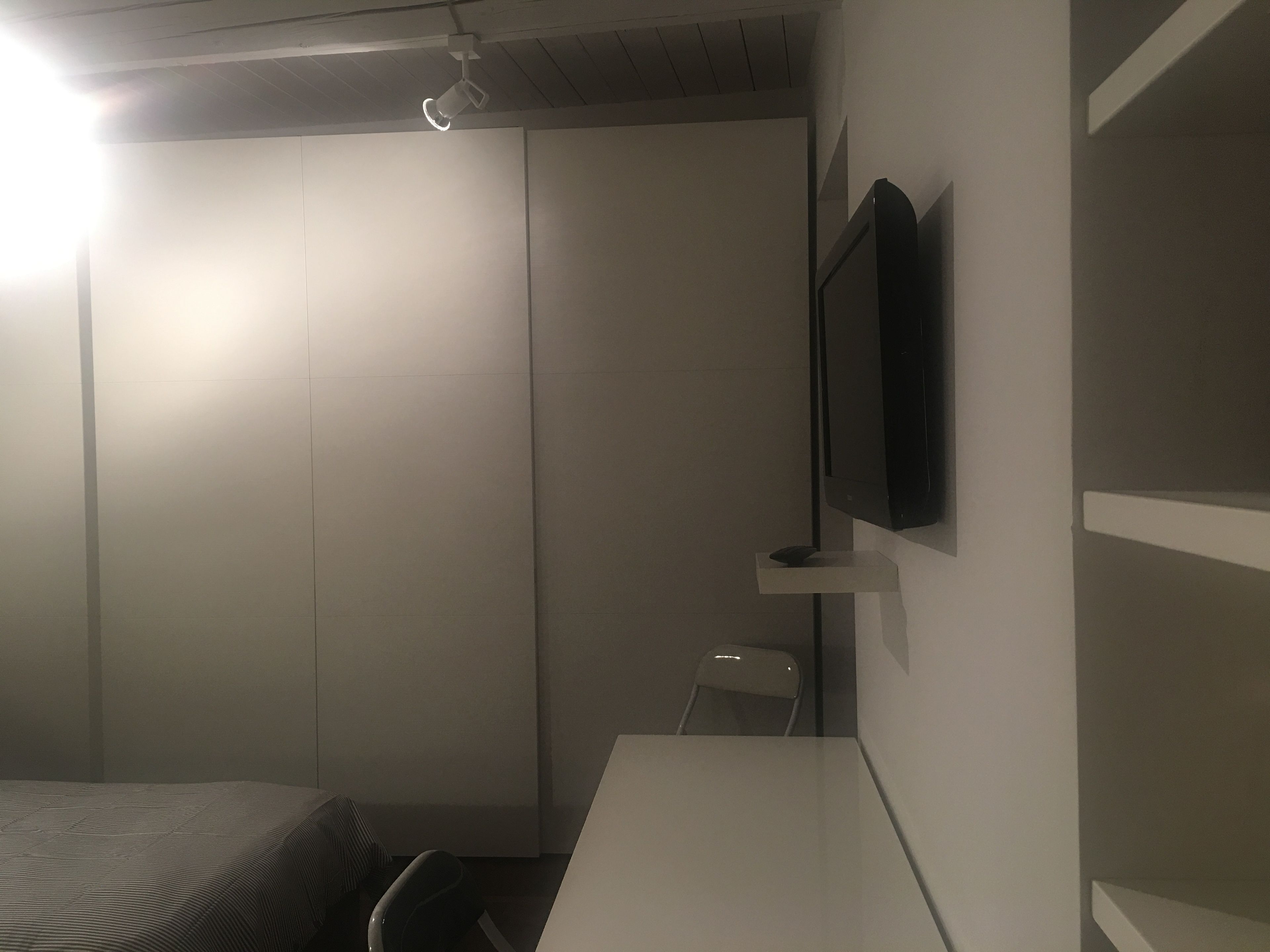 A room with a bed, a desk, a chair, a television mounted on the wall, and a ceiling light.