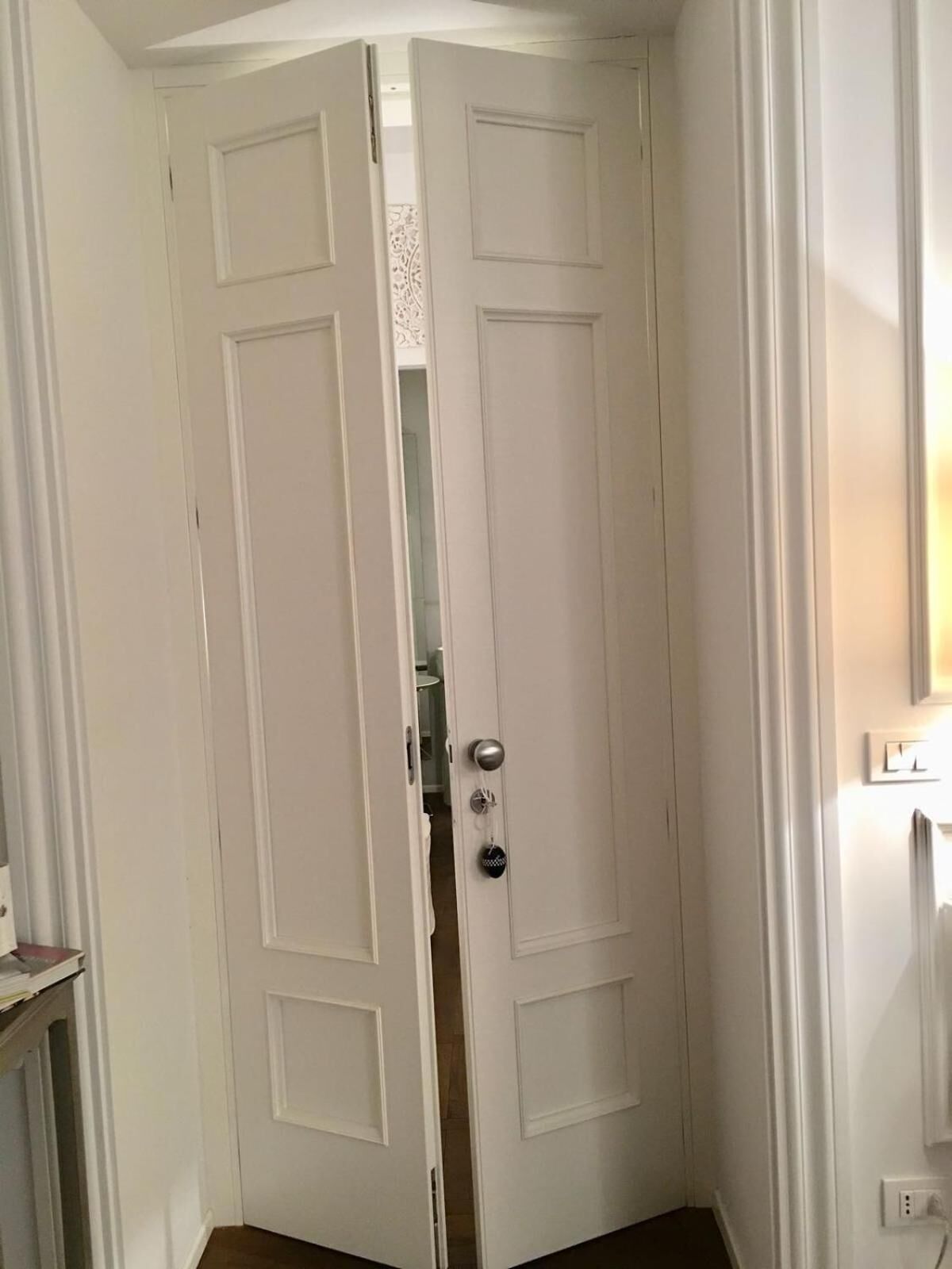 A white door with a silver handle and a keyhole, leading to a room with a white wall and a small window.