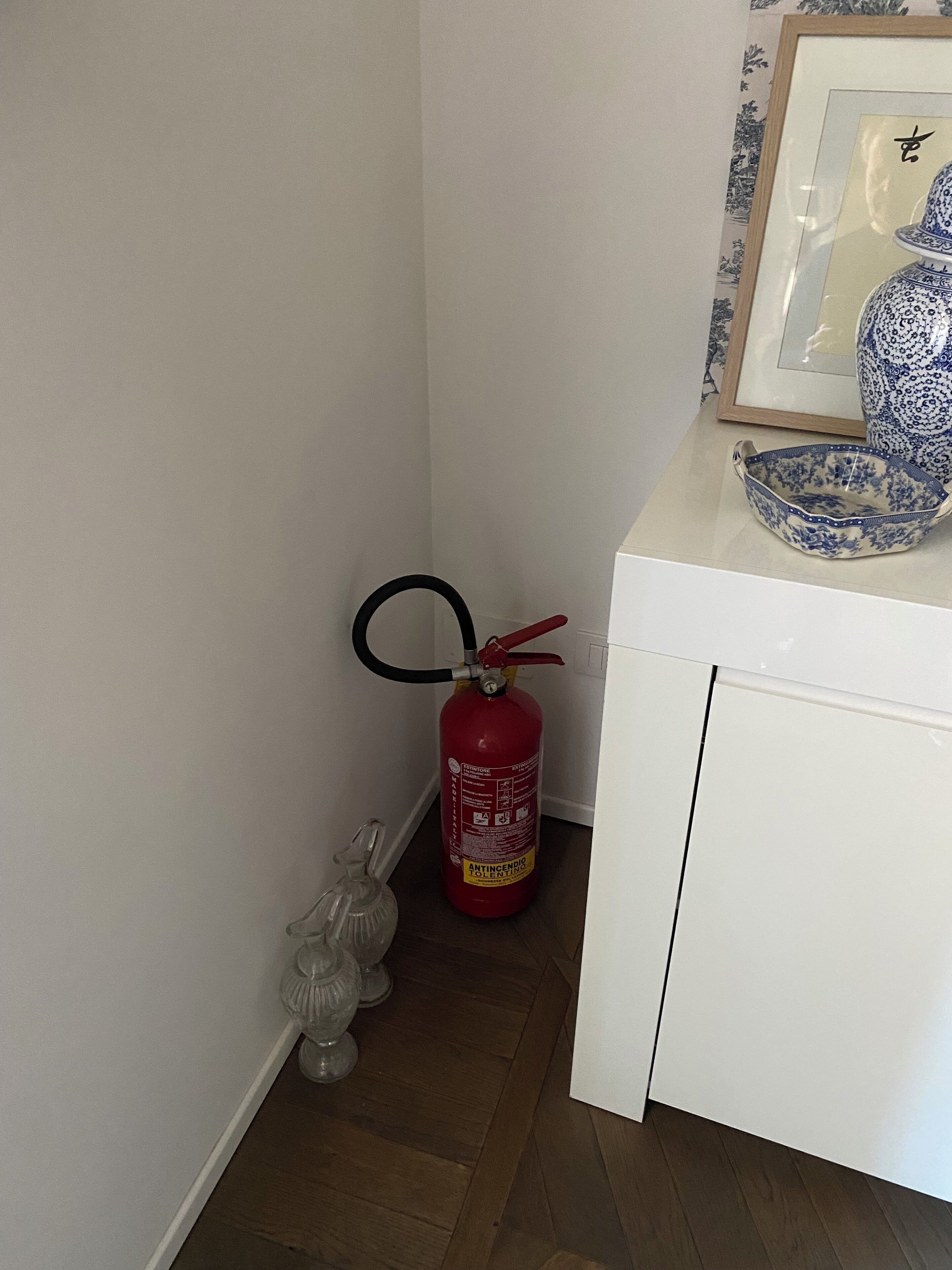 Un armario blanco con un jarrón azul y blanco enmarcado encima. Un extintor rojo está colgado en la pared junto al armario.