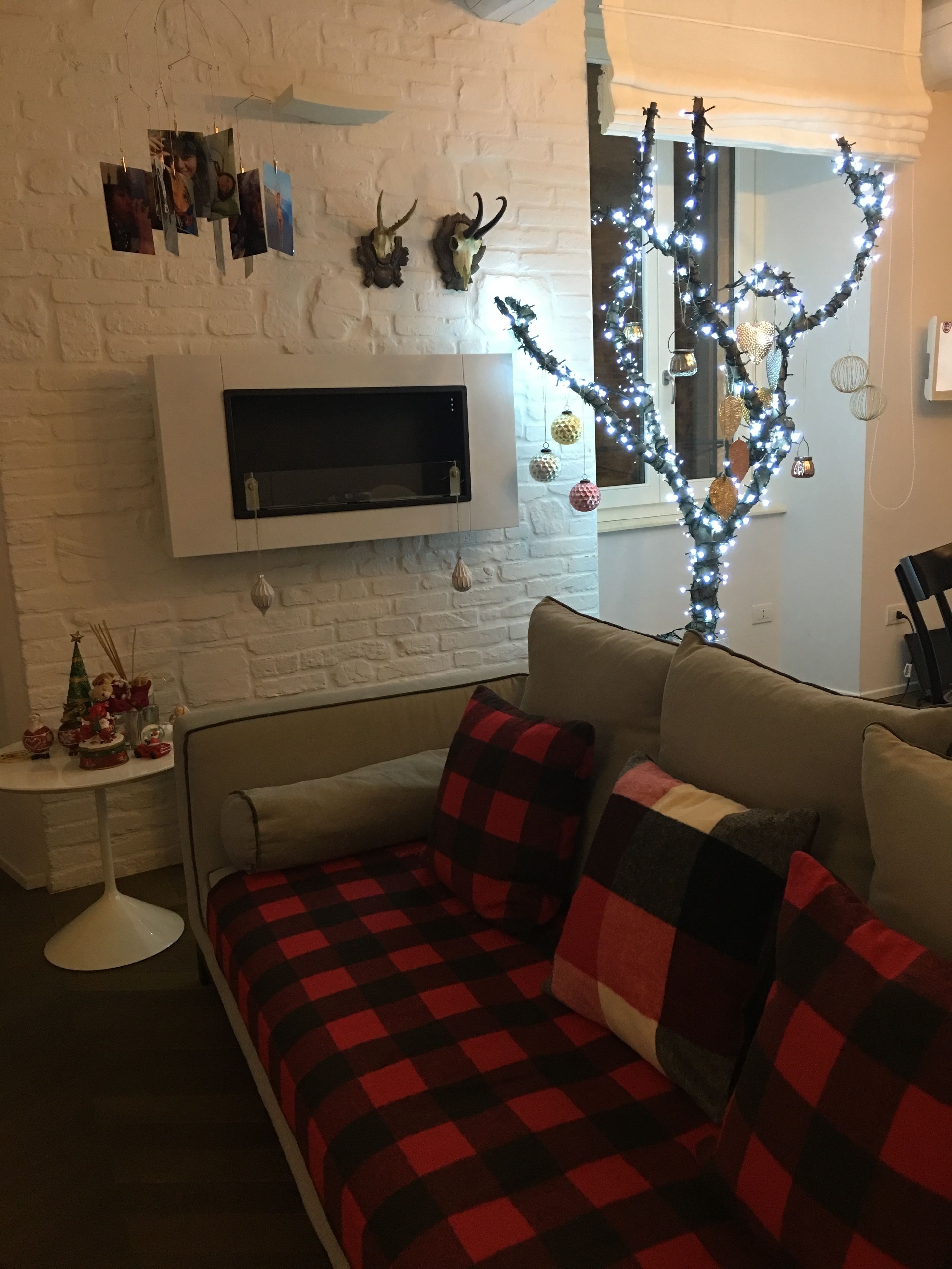 A cozy living room with a sofa, a small side table, a white brick wall, a mounted TV, and a decorative tree with lights.