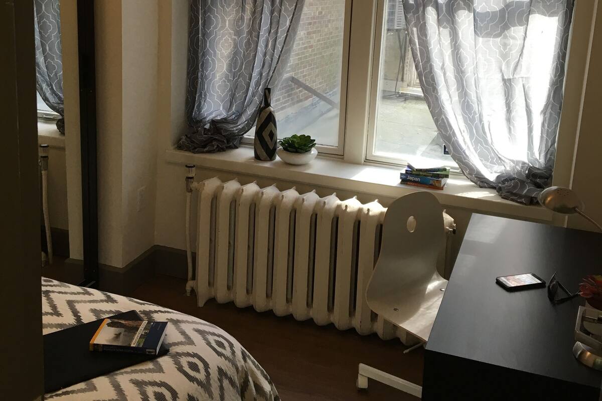 A room with a bed, a desk, a chair, a radiator, and a window with curtains.
