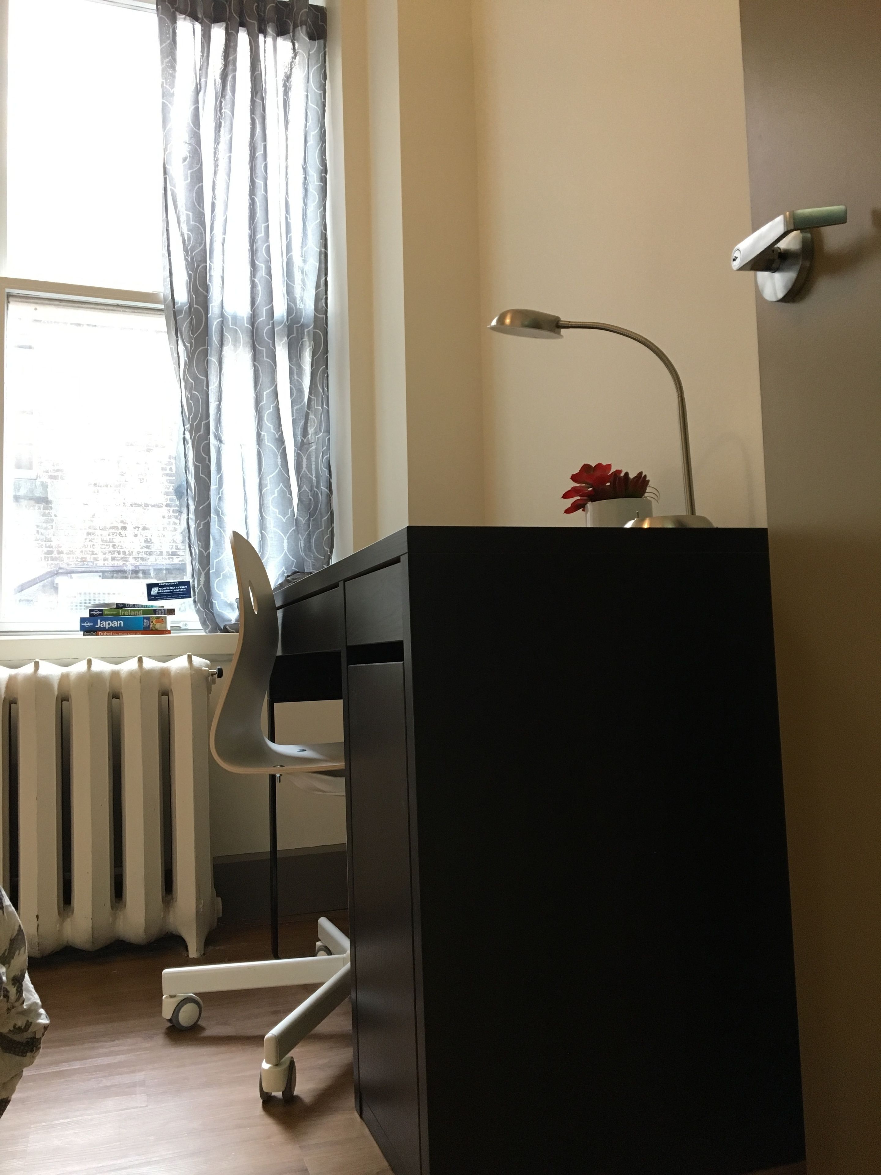 Une pièce comprenant un bureau, une chaise, un radiateur, une fenêtre avec des rideaux et une lampe.