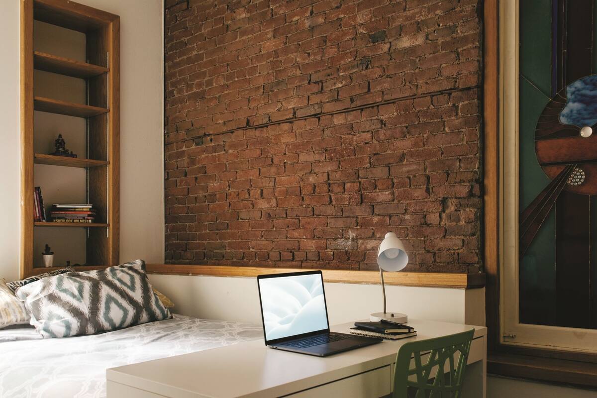 A bedroom with a brick wall, a bed with patterned pillows, a desk with a laptop, a green chair, and a hanging light fixture.