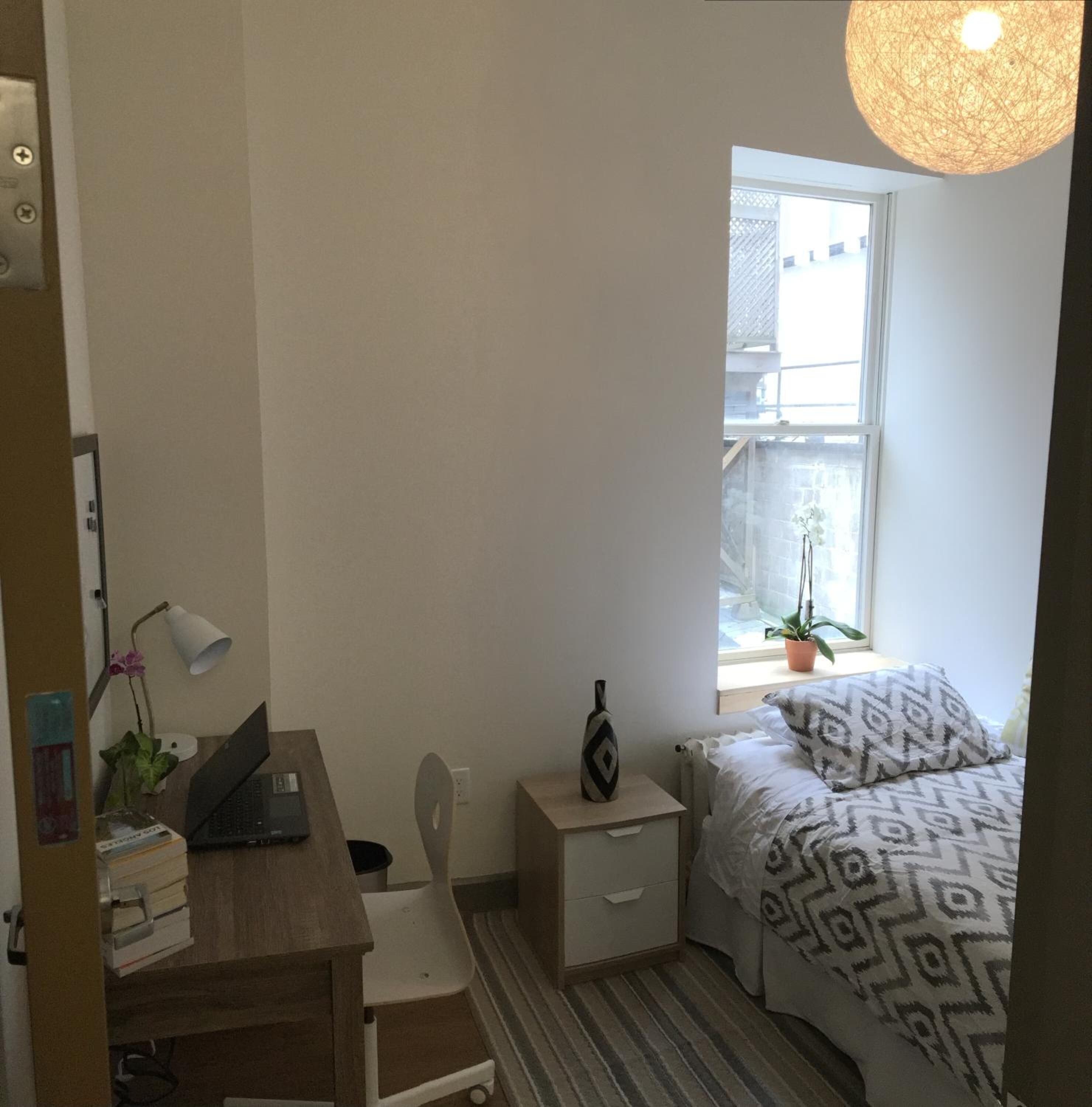 Une petite chambre à coucher, bien rangée, avec un lit, un bureau, une chaise, une table de chevet, une lampe et une fenêtre donnant sur l’extérieur.