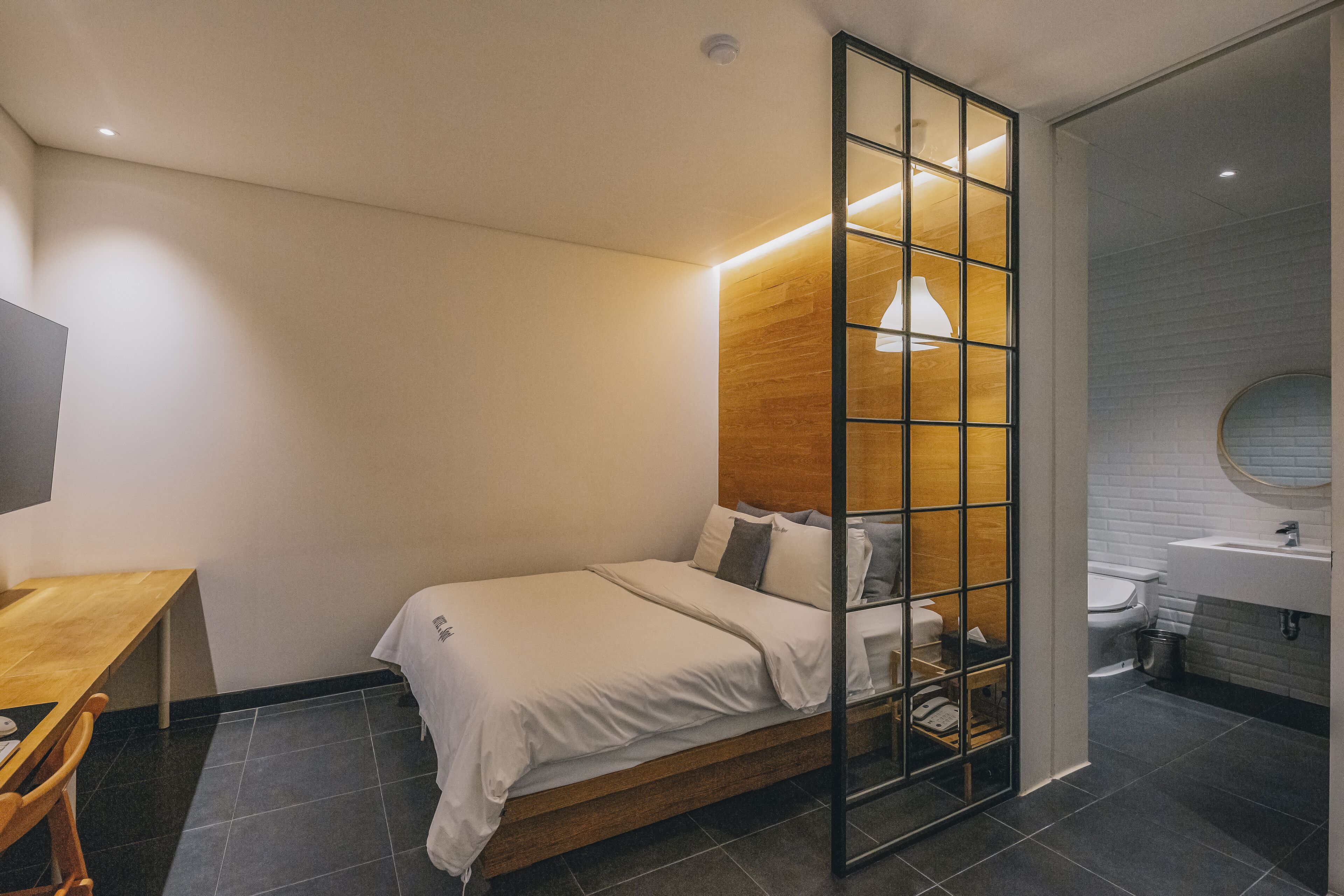 A modern bedroom with a wooden headboard, a bed with white bedding, a desk with a chair, and a bathroom with a sink and toilet.