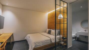 A modern bedroom with a wooden headboard, a bed with white bedding, a desk with a chair, and a bathroom with a sink and toilet.