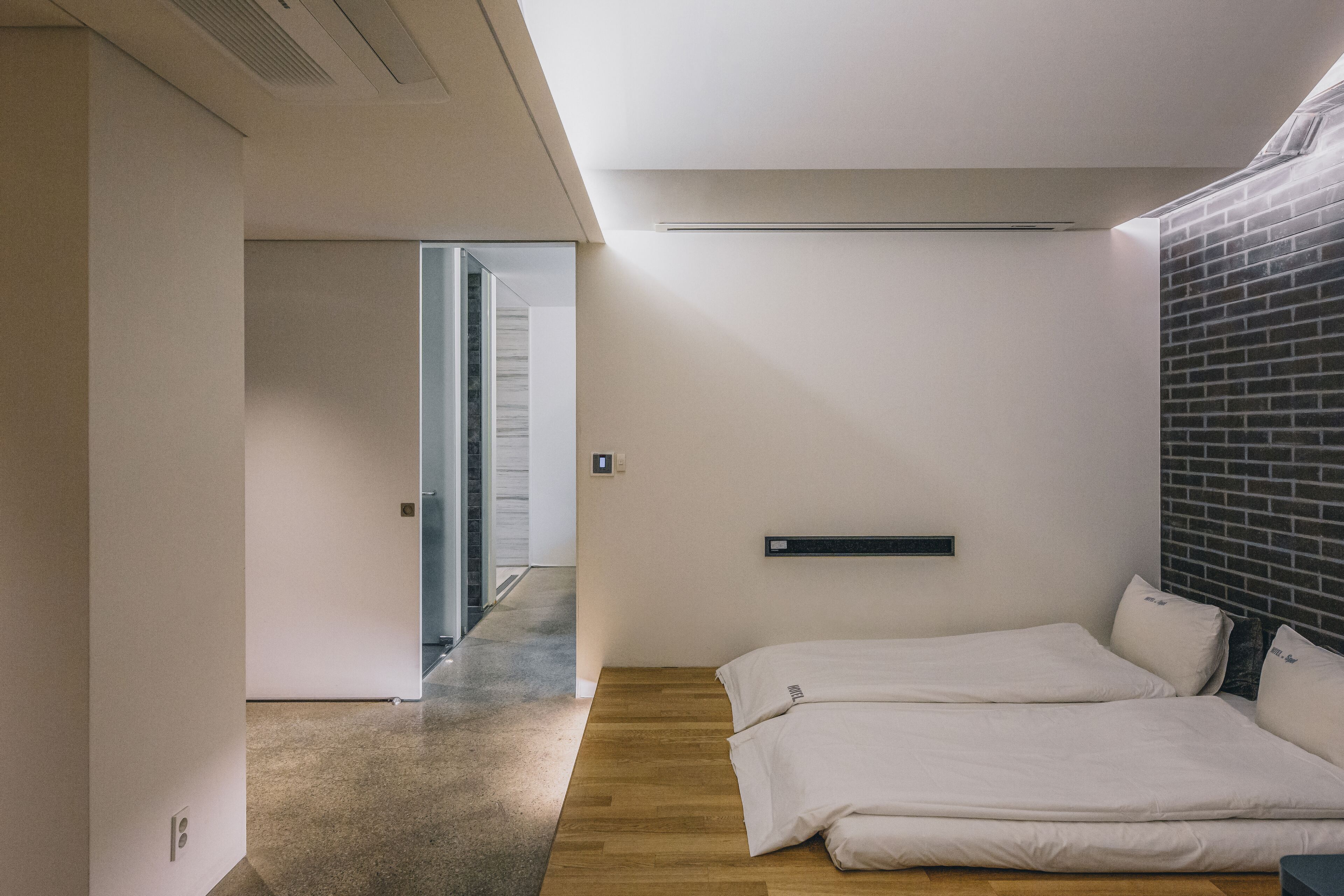 A modern hotel room with a wooden floor, two beds, a brick accent wall, and a minimalist design.