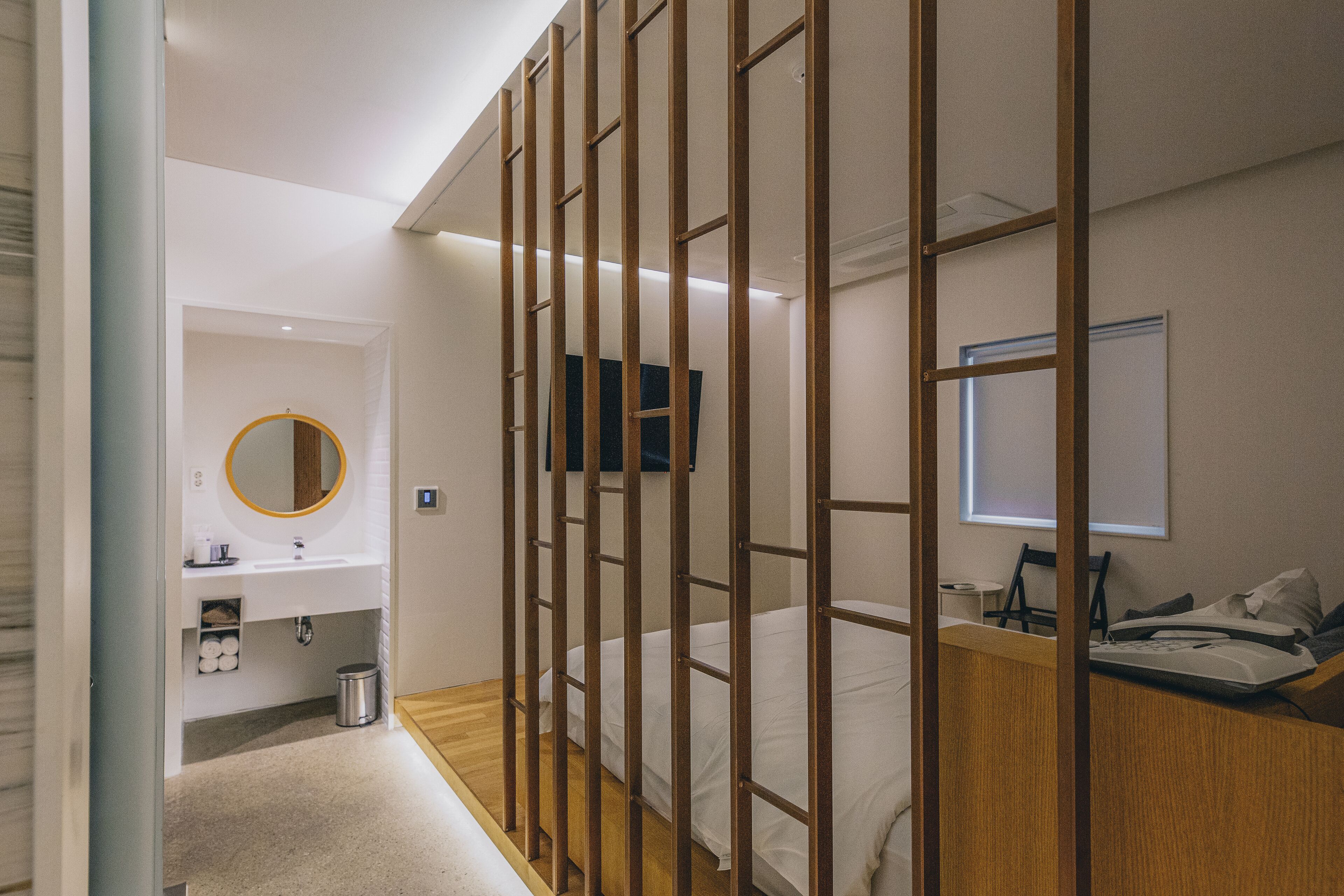 A modern hotel room with a wooden partition, a bed, a desk with a chair, a bedside table, a mirror, and a window.