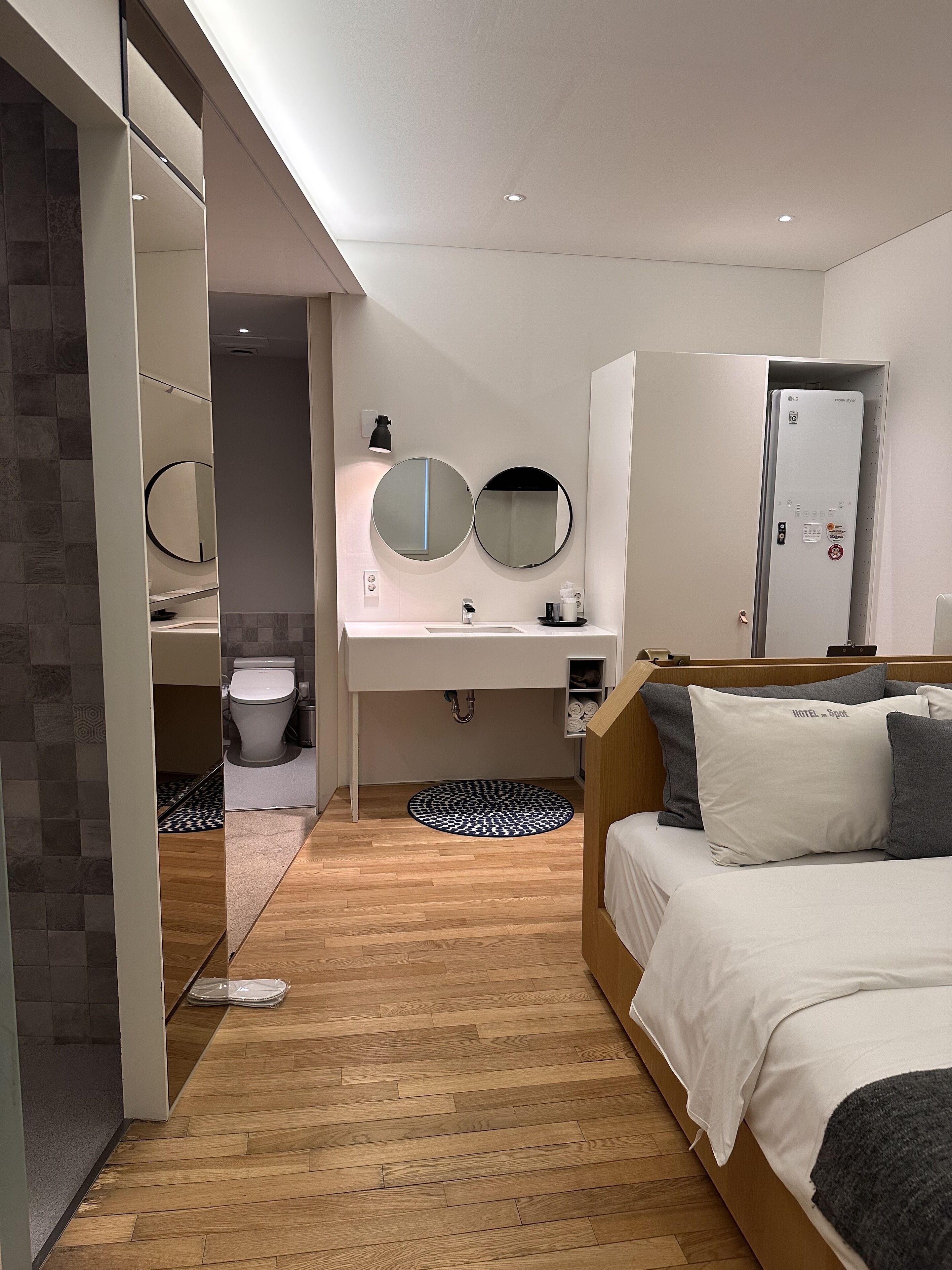 Una habitación de hotel moderna con piso de madera, una cama con ropa de cama blanca y gris, un espejo redondo sobre el lavamanos y un armario empotrado.
