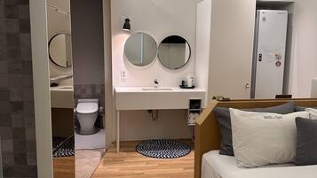 Una habitación de hotel moderna con piso de madera, una cama con ropa de cama blanca y gris, un espejo redondo sobre el lavamanos y un armario empotrado.