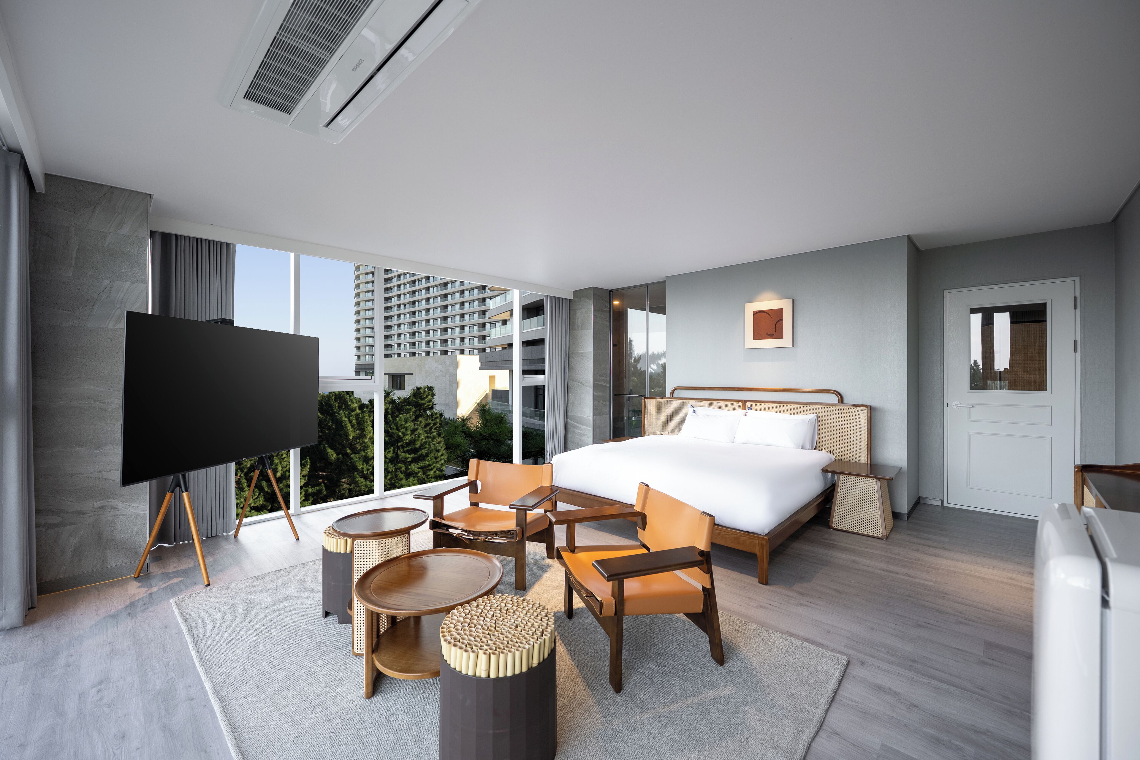 A modern hotel room with a large bed, a flat-screen TV, a seating area with chairs and a round table, and a view of the city through large windows.