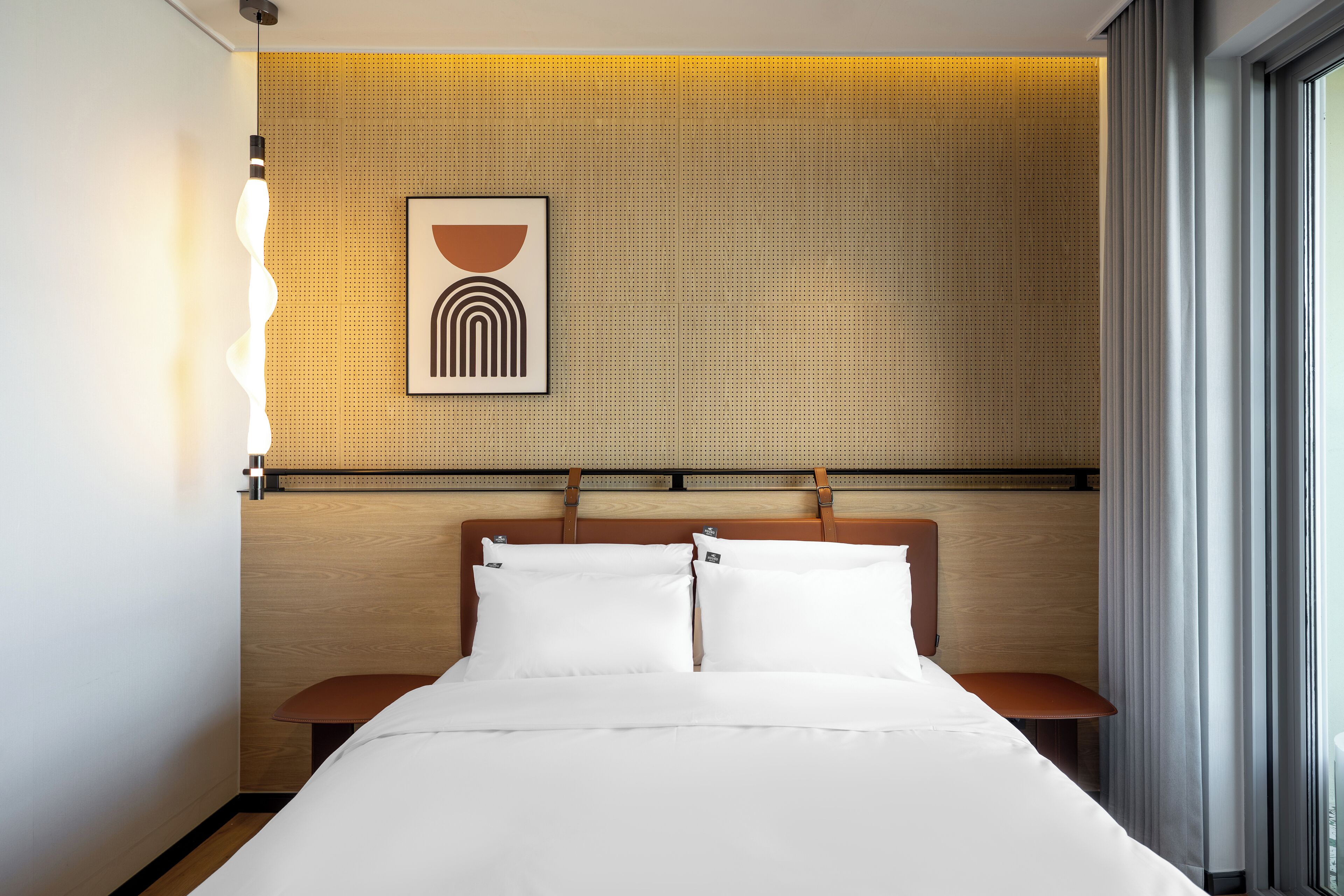 A hotel room with a bed, a wooden headboard, a wall-mounted artwork, and a window with curtains.