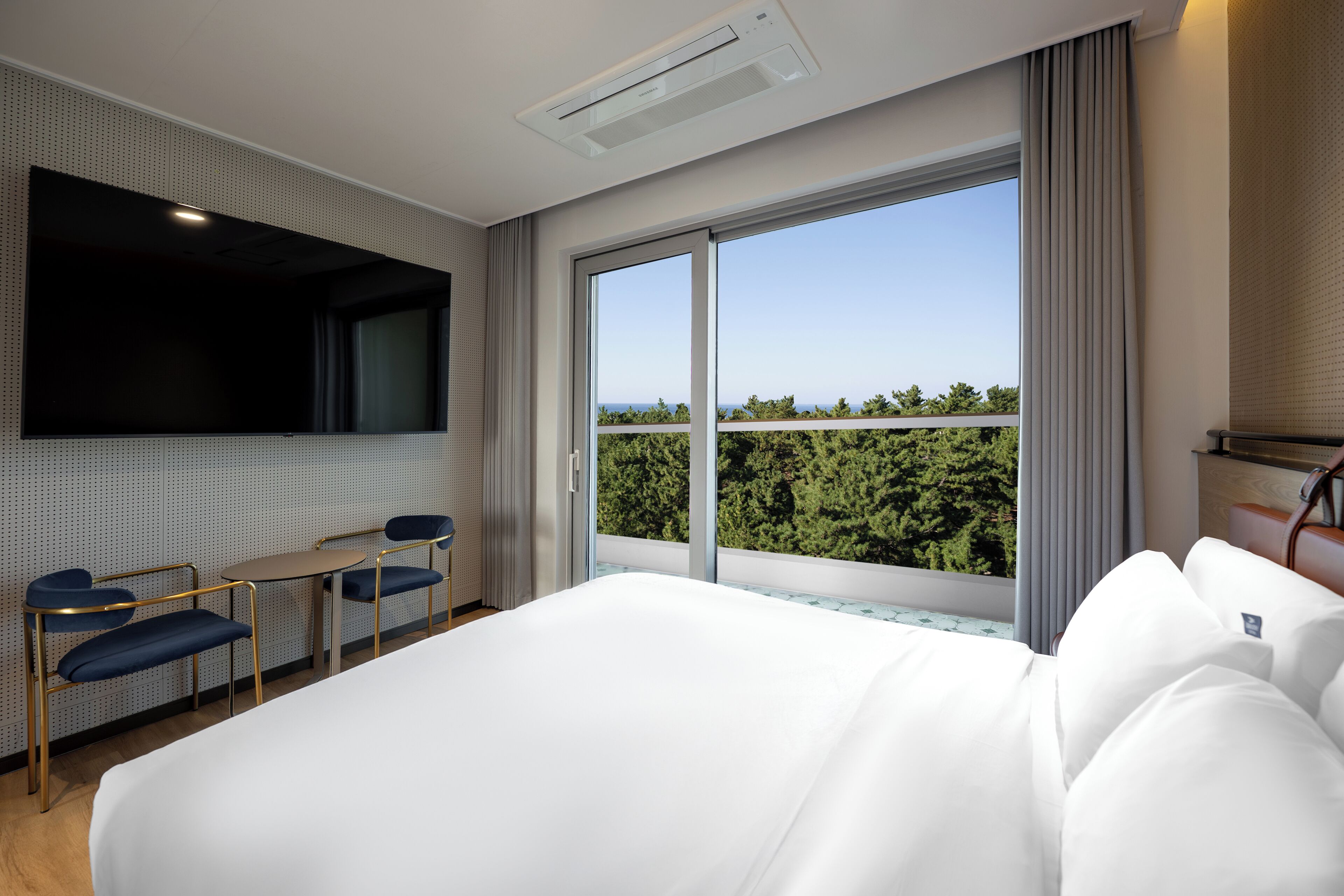 A hotel room with a large bed, a desk with two chairs, a flat-screen TV, and a balcony with a view of trees.