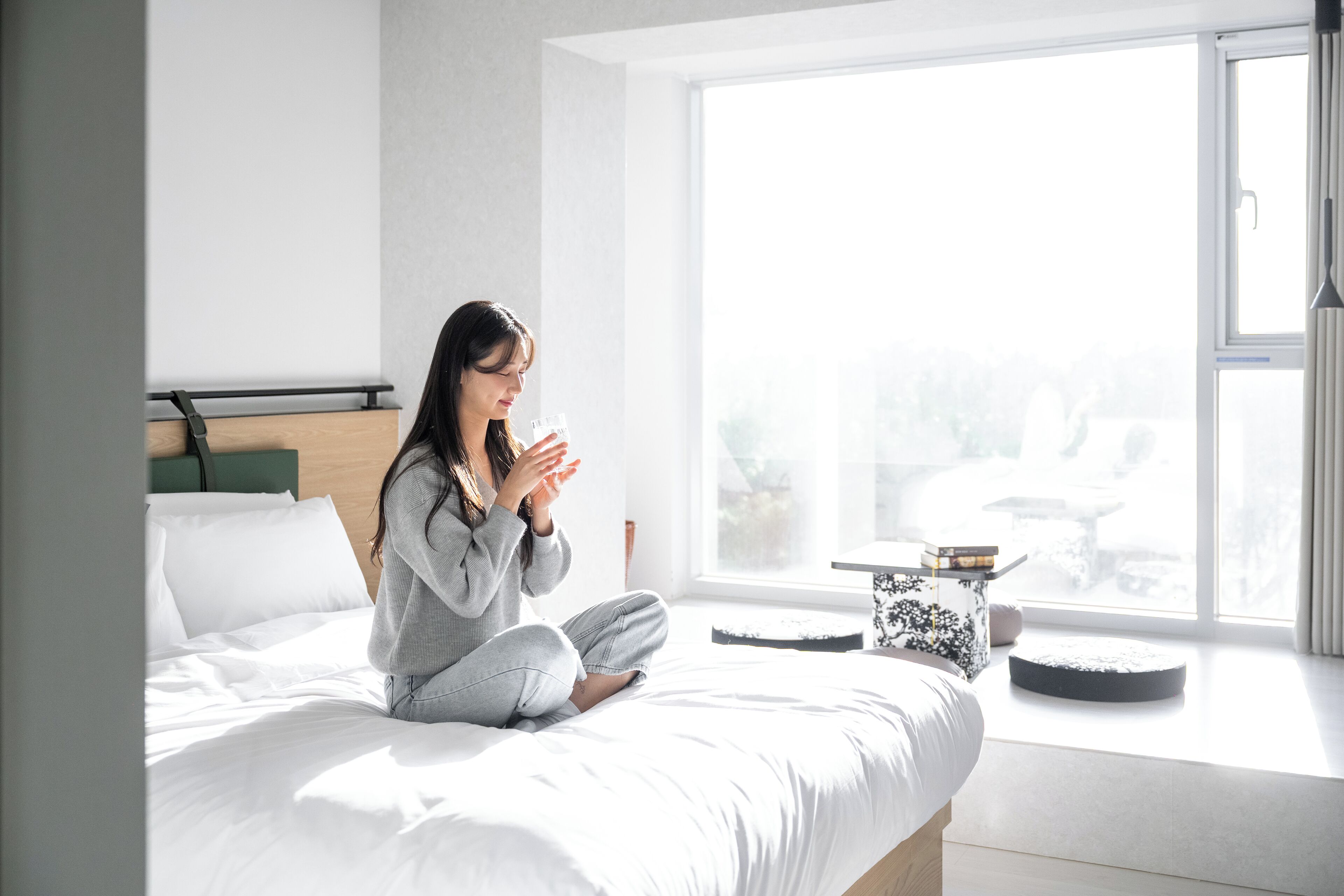 A person sitting on a bed in a room with a large window, a bedside table, and a view of the outdoors.