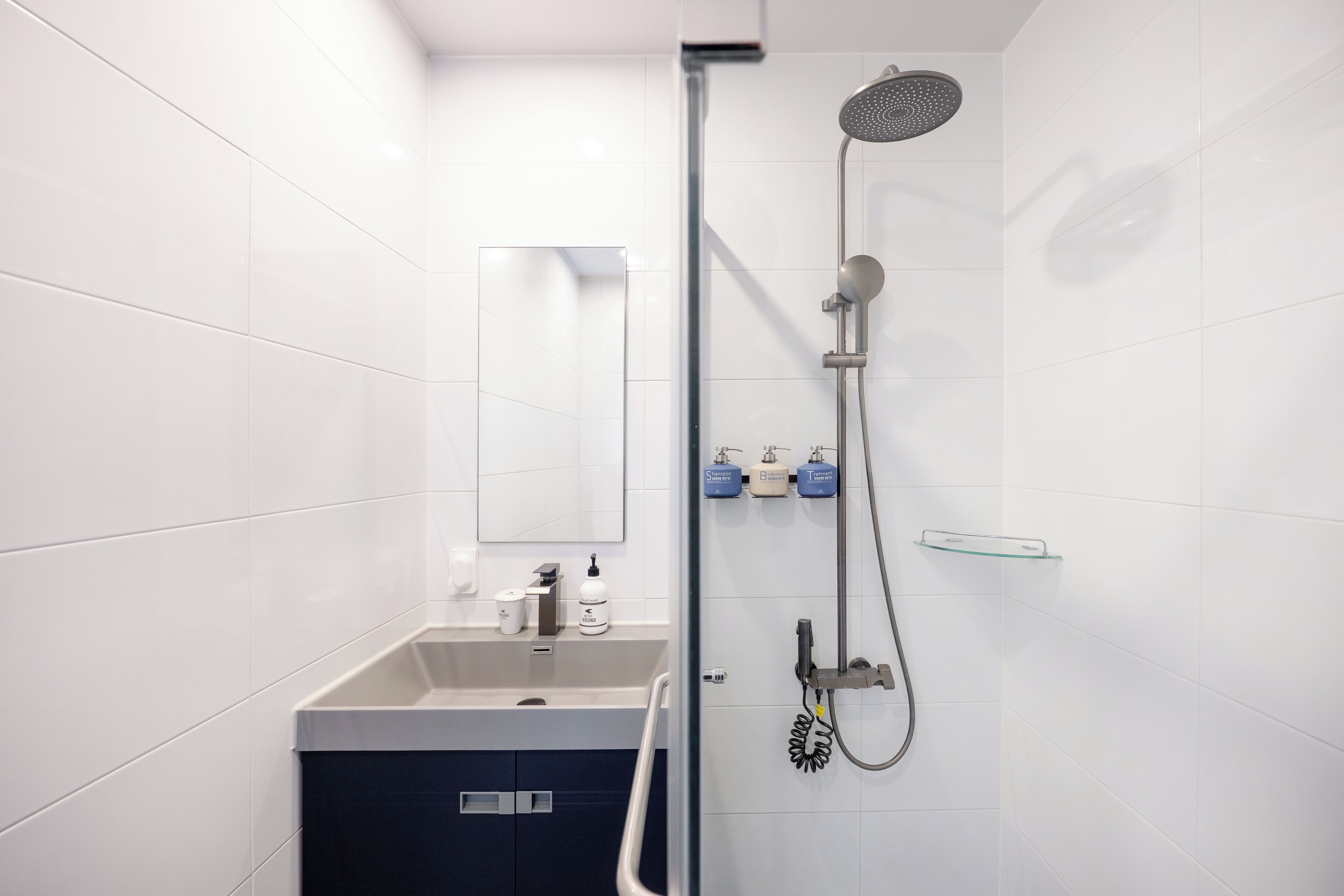 A modern bathroom with white tiles, a glass shower enclosure, a sink with a mirror, and toiletries on the wall.