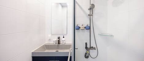 A modern bathroom with white tiles, a glass shower enclosure, a sink with a mirror, and toiletries on the wall.