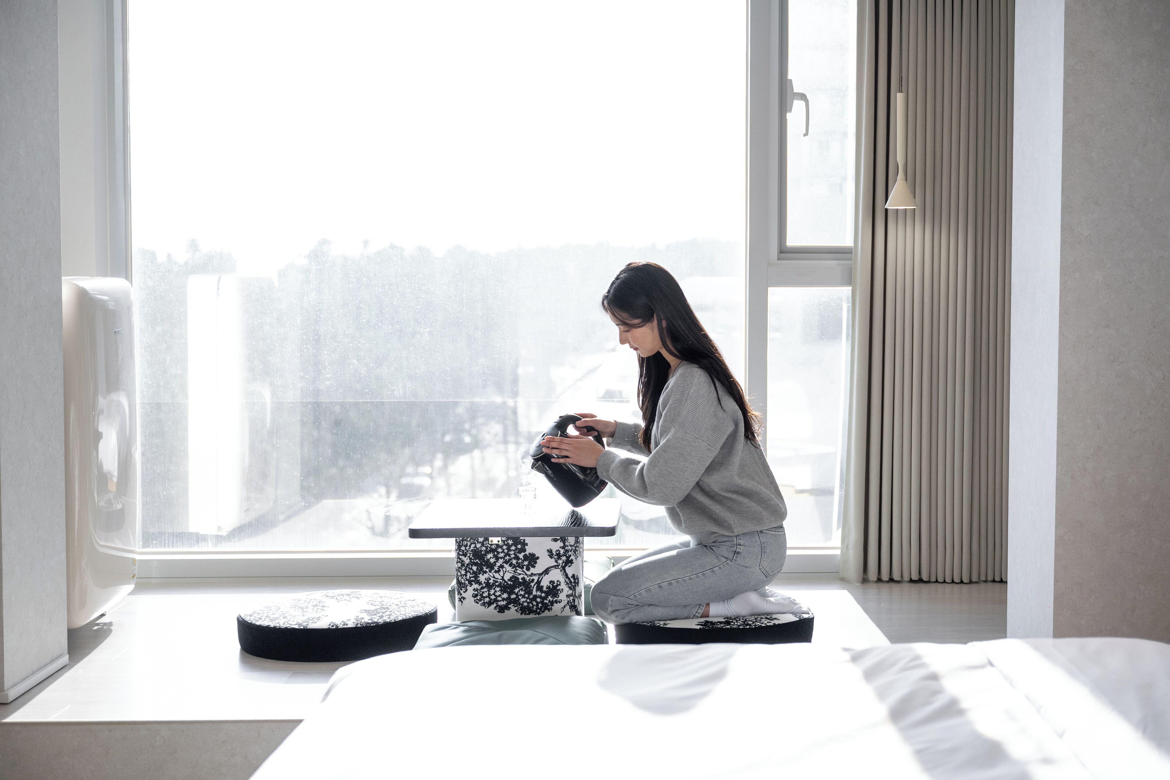 A person sitting on a bed in a room with a large window, a bedside table, and a lamp.