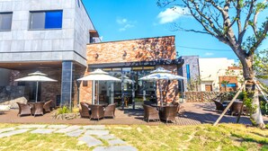 An outdoor seating area with wicker chairs and umbrellas, a stone pathway, and a modern building with glass doors. - Marvillrick Spa&Poolvilla (Jeju City)