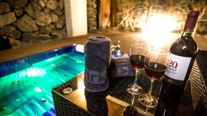 A hot tub with a stone wall backdrop, two wine glasses, and a bottle of wine on a table.