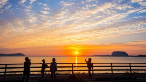 A sunset over a body of water with a distant landmass, viewed from a waterfront promenade with a railing.