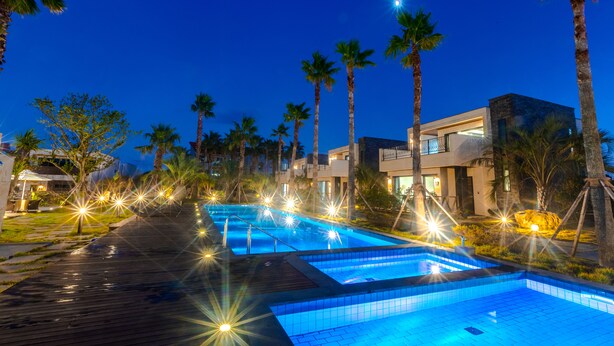 A swimming pool with a wooden deck, surrounded by palm trees and modern buildings. - Marvillrick Spa&Poolvilla (Jeju City)