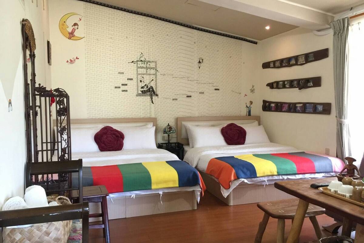 A room with two beds, a wooden bench, a small table with cups, and a wall with a decorative pattern and framed photos.