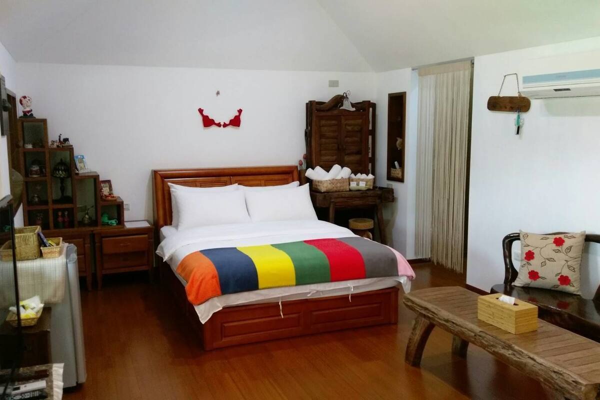 A bedroom with a wooden bed, a bedside table, a chair, a small table with a tissue box, and a window with curtains.