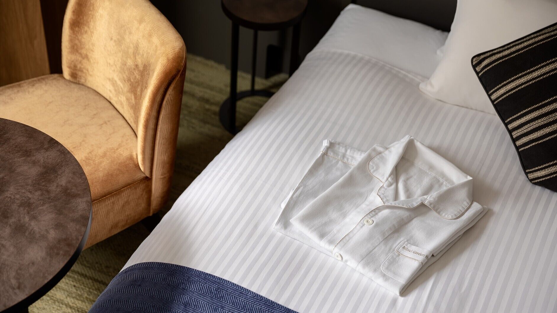 A neatly made bed with a folded white shirt on it, a striped pillow, and a brown chair in the room.