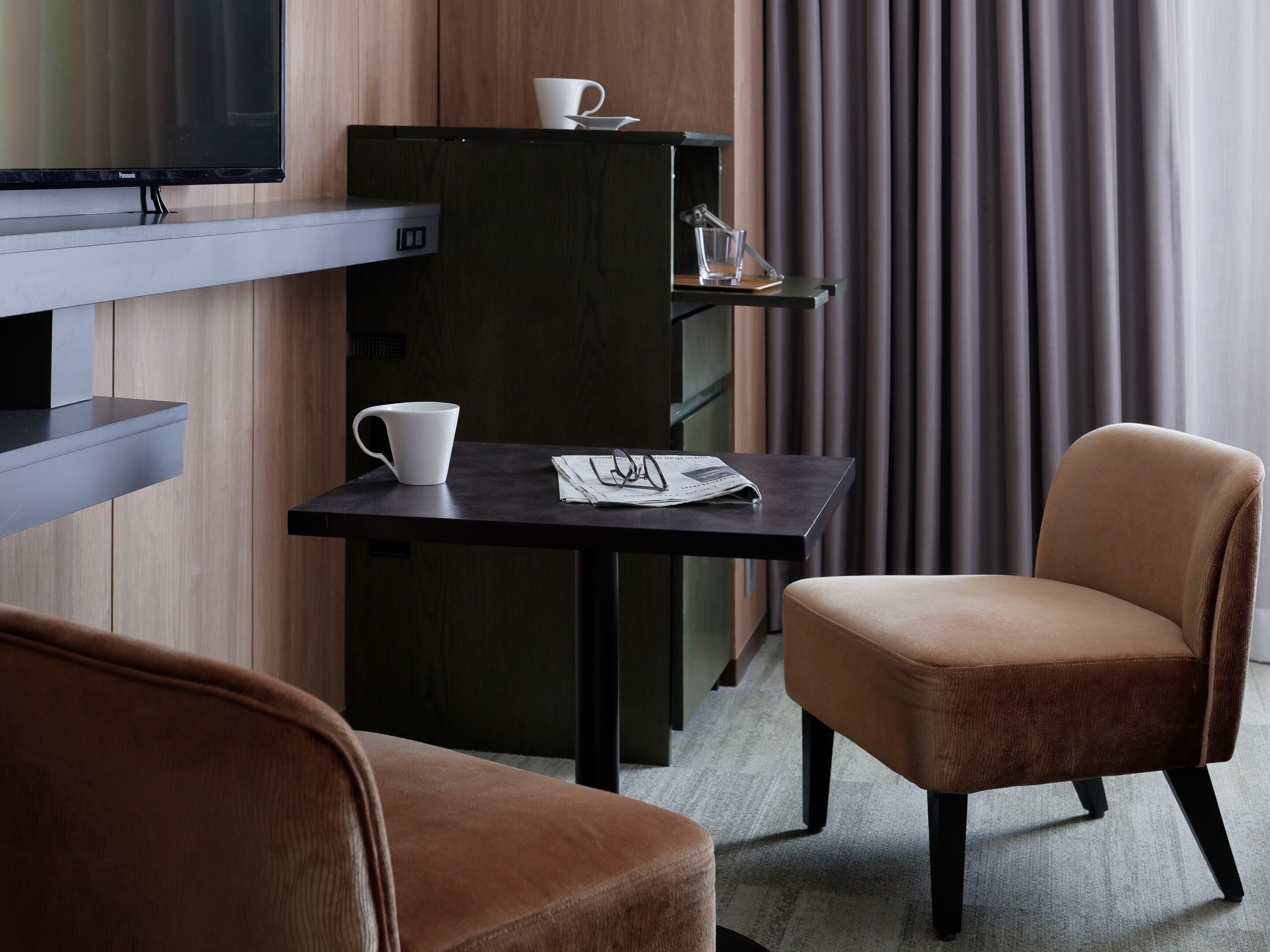 A hotel room with a flat-screen TV, a small table with a cup and glasses, a chair, and a cabinet with a cup on top.