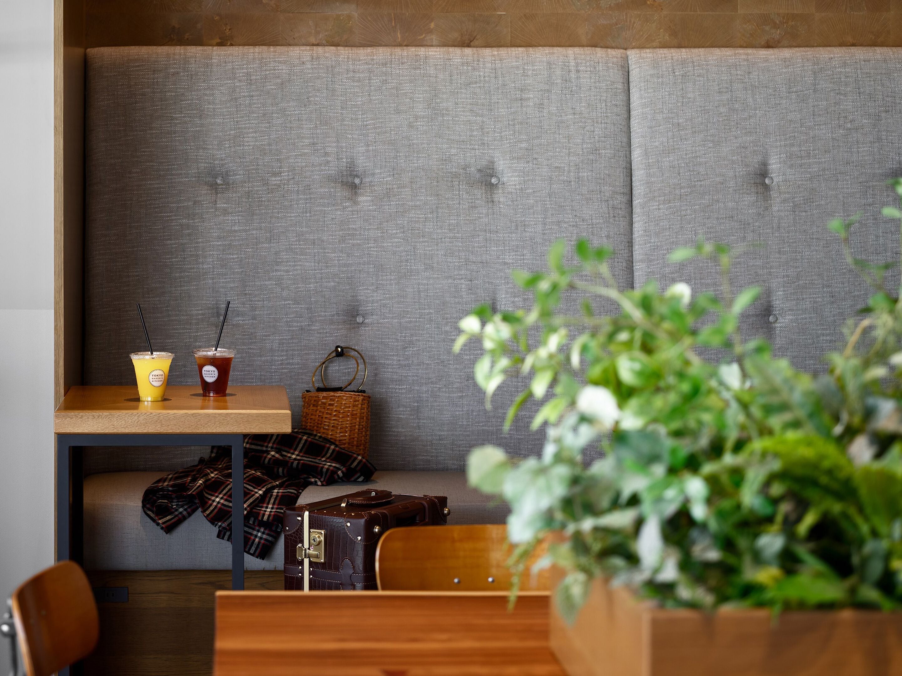A seating area with a grey upholstered bench, a wooden table with two drinks, a wicker basket, and a plaid blanket.