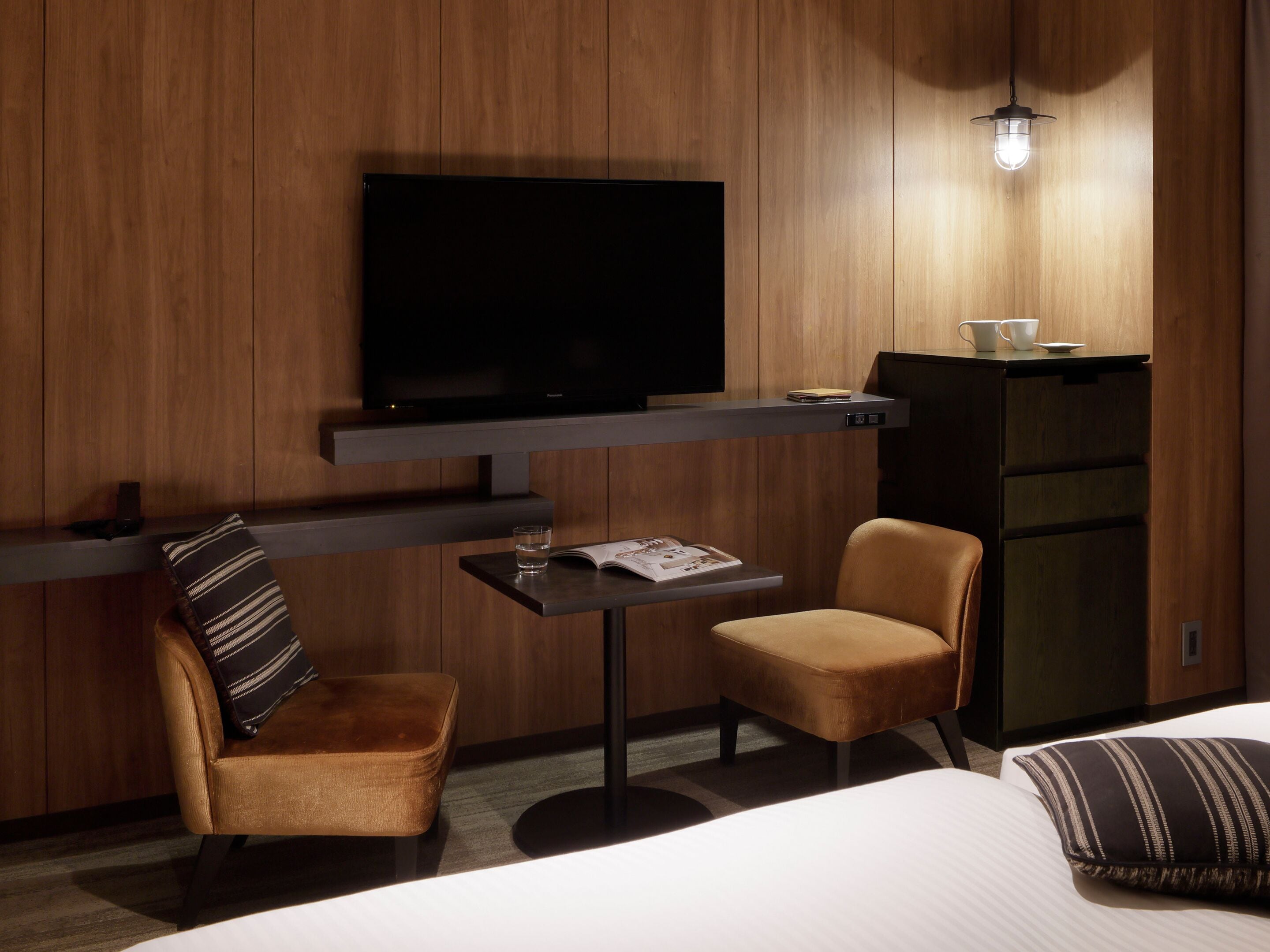 A hotel room with a TV mounted on the wall, a small table with a glass of water, a cushioned chair, and a bed with a striped pillow.