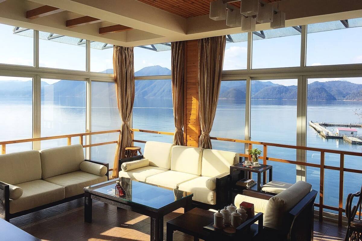 Quarto amplo com grande janela que oferece vista para um lago e montanhas. Há um sofá, uma mesa de centro e uma mesinha com bules de chá.