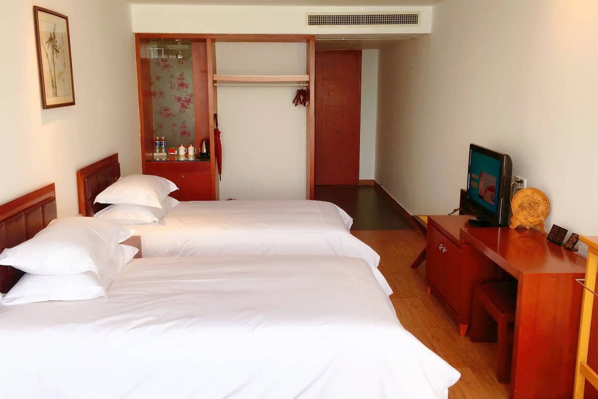 Quarto de hotel com duas camas, uma cômoda de madeira, uma mesa de escritório com cadeira, uma televisão e uma mesinha com um objeto decorativo.