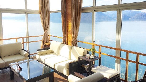 A spacious room with a large window overlooking a calm lake and mountains. There is a white sofa, a glass coffee table, and a small side table with a vase and books.