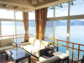 Une pièce spacieuse dotée d’une grande fenêtre donnant sur un lac paisible et des montagnes. On y trouve un canapé blanc, une table basse en verre et une petite table d’appoint avec un vase et des livres.