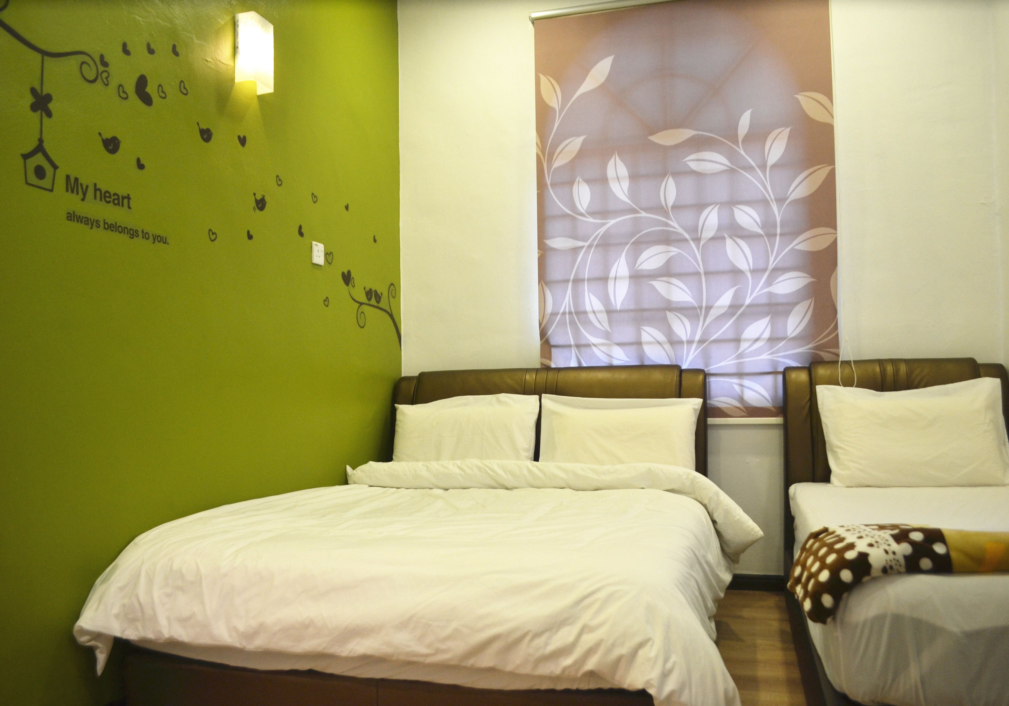A hotel room with two beds, a green wall with a heart decoration, and a large window with a leaf pattern.