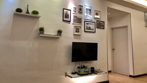 A modern living room with a flat-screen TV mounted on a shelf, surrounded by framed photographs and small potted plants. - H & H Guesthouse 1 (Malacca City)