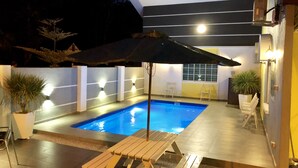 A swimming pool with a wooden lounge chair and an umbrella, surrounded by walls with light fixtures and potted plants. - H & H Guesthouse 1 (Malacca City)