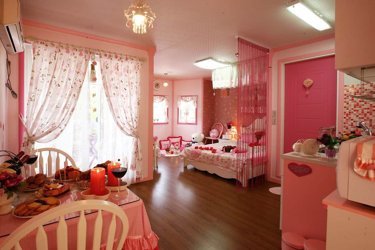Quarto em tons de rosa, com mesa de jantar posta para refeição, cama com roupa de cama rosa e porta rosa.