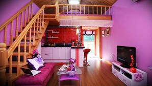 Una sala de estar acogedora con un sofá morado, una escalera de madera, una cocina con baldosas rojas y un televisor sobre un mueble blanco.