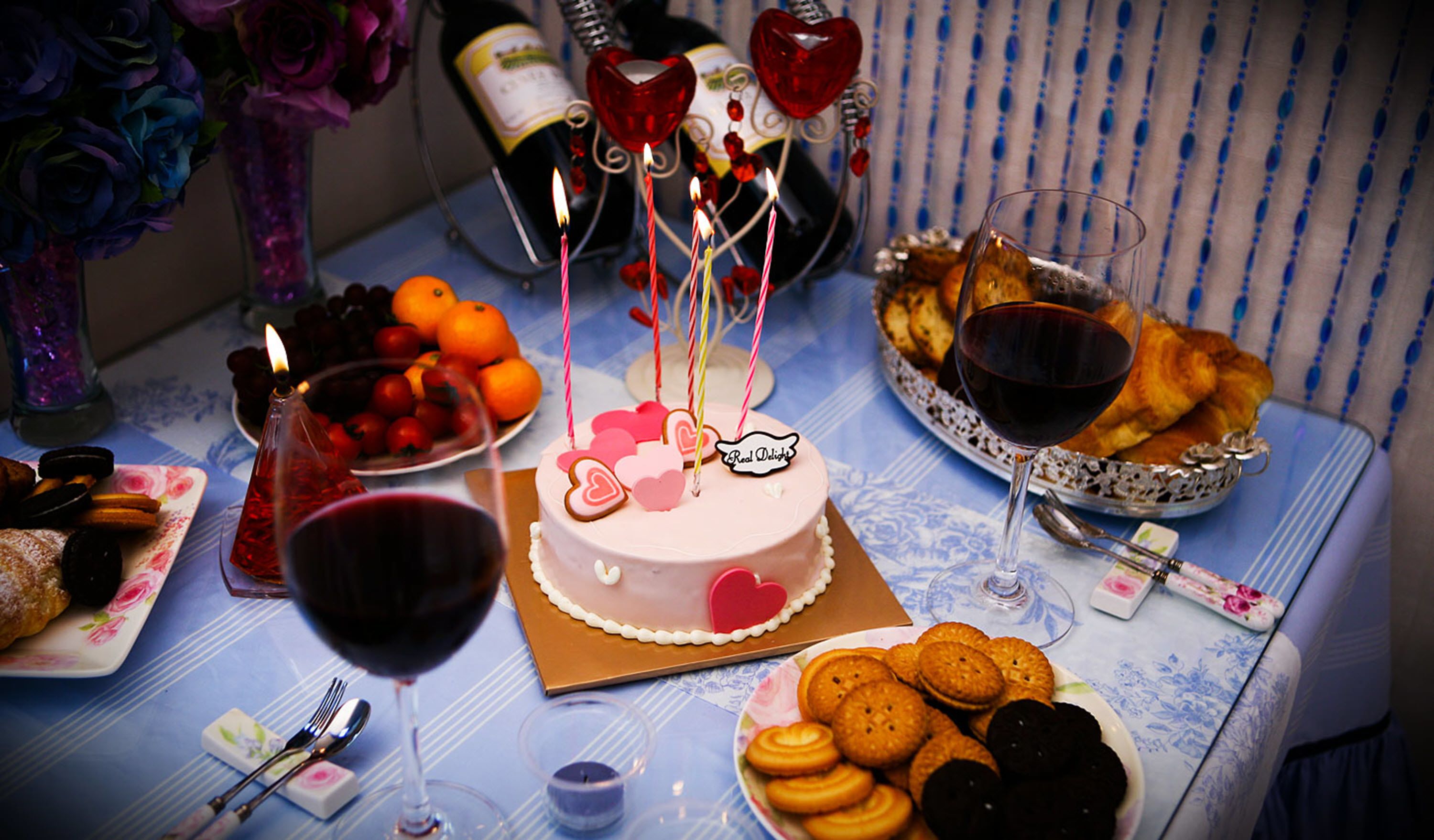 Mesa posta com bolo, taças de vinho, frutas e doces, indicando uma ocasião festiva.