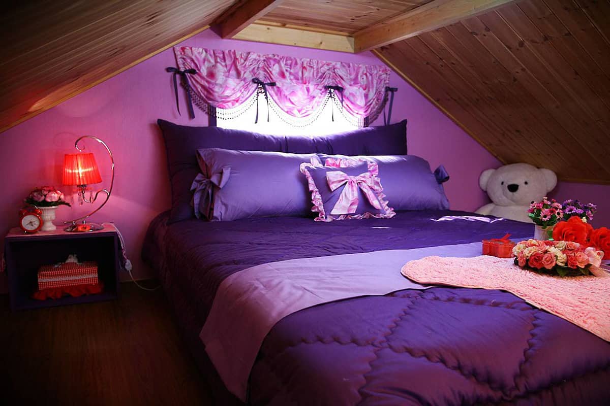 Quarto com teto inclinado, roupa de cama roxa, um urso de pelúcia e uma mesinha de cabeceira com abajur e flores.