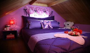 Une chambre avec un plafond en pente, une literie violette, un ours en peluche et une table de chevet avec une lampe et des fleurs.