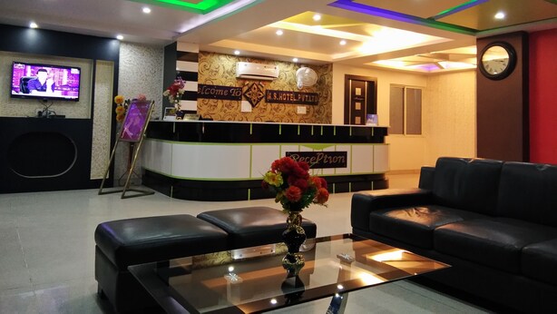 A hotel reception area with a modern design, featuring a reception desk, a television, a clock, and a seating area with a coffee table and a vase of flowers. - M S Hotel (Bhagalpur)