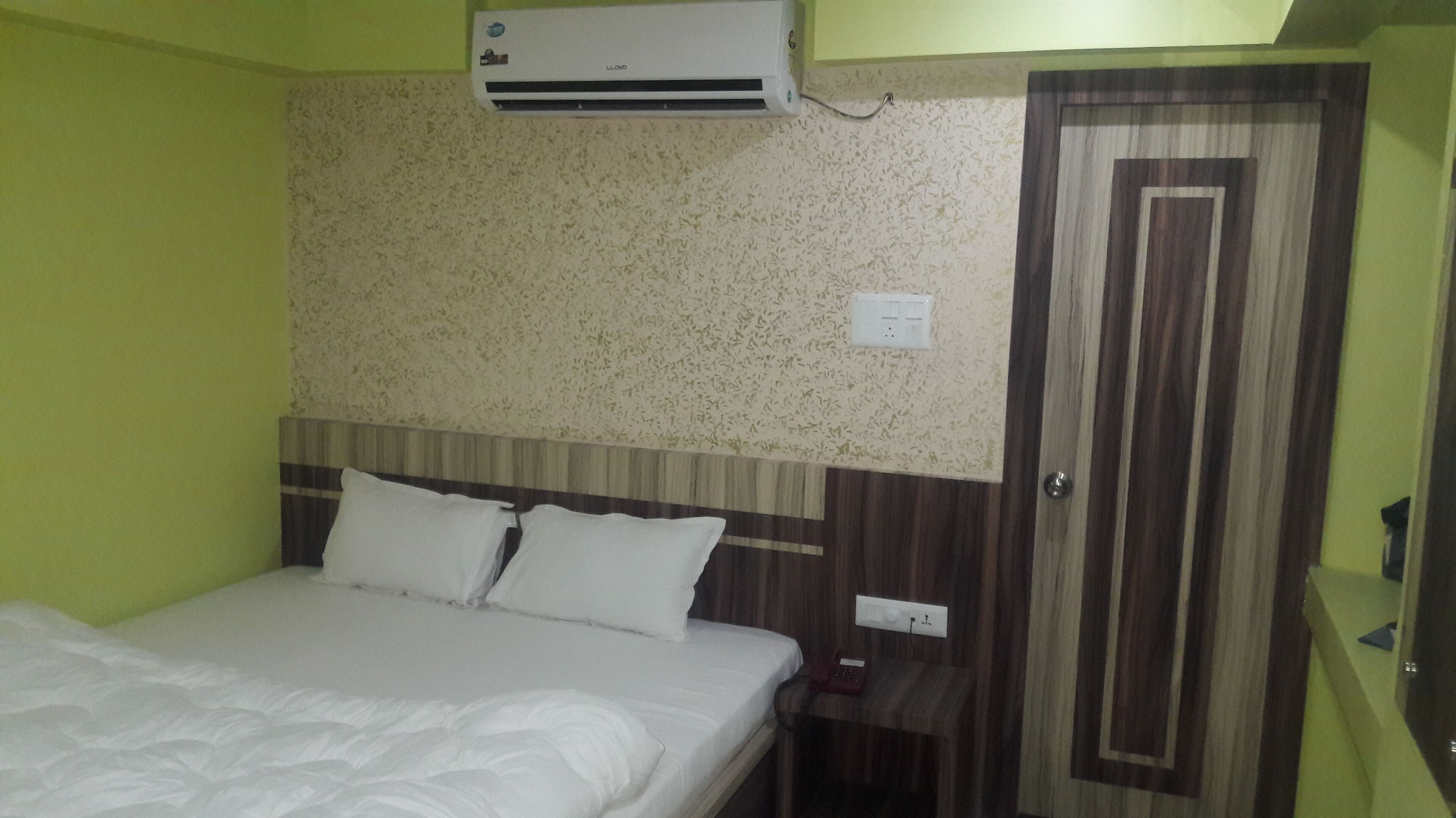 A hotel room with a bed, a nightstand, a wall-mounted air conditioner, and a door.